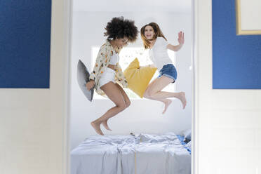 Carefree female friends holding pillow while jumping on bed at home - JCCMF03270