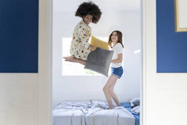 Young female friends with pillow jumping on bed at home - JCCMF03272
