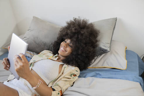 Lächelnde junge Frau, die ein digitales Tablet benutzt, während sie zu Hause auf dem Bett liegt - JCCMF03278