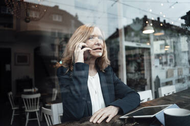 Senior blond businesswoman talking on smart phone sitting in cafe seen through glass window - JOSEF05093