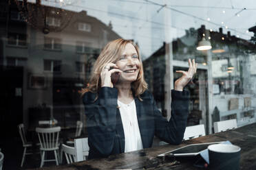 Glückliche weibliche Fachkraft, die in einem Café sitzt und mit einem Mobiltelefon spricht, gesehen durch ein Glasfenster - JOSEF05095