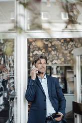 Mature businessman with hand in pocket talking on smart phone standing at doorway - JOSEF05097