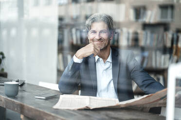 Smiling businessman with newspaper sitting in cafe seen through glass window - JOSEF05109