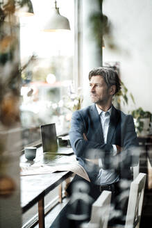 Männlicher Berufstätiger mit verschränkten Armen, der durch das Fenster schaut, während er in einem Cafe sitzt - JOSEF05120