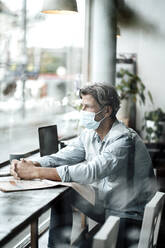 Mature man sitting with hands clasped looking through window while sitting in cafe during COVID-19 - JOSEF05121