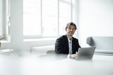 Mature businessman sitting with laptop at desk in office - JOSEF05125