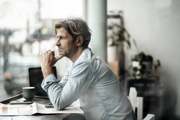 Thoughtful man sitting with newspaper looking through window while sitting in cafe - JOSEF05127