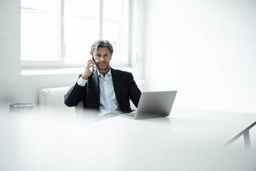 Männlicher Berufstätiger sitzt mit Laptop und spricht mit seinem Smartphone im Büro - JOSEF05128