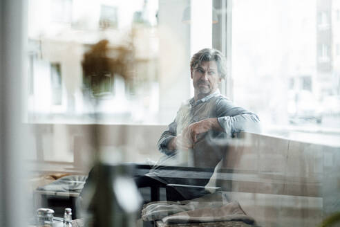 Reifer Mann schaut weg, während er in einem Café sitzt, gesehen durch ein Glasfenster - JOSEF05138
