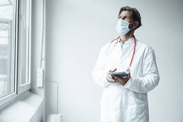 Male doctor wearing protective face mask holding digital tablet while standing by window in clinic - JOSEF05150