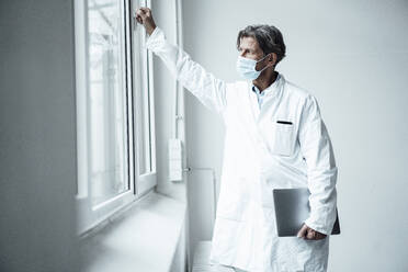 Reifer männlicher Arzt mit Gesichtsschutzmaske schaut durch ein Fenster im Krankenhaus - JOSEF05153