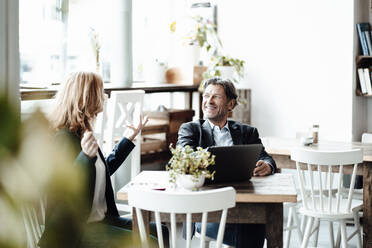 Happy businessman with laptop looking at female professional discussing while sitting in cafe - JOSEF05160