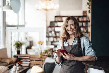 Lächelnde Café-Besitzerin, die ihr Smartphone hält, während sie in einem Café steht - JOSEF05170