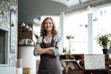 Happy blond female cafe owner wearing apron standing with arms crossed - JOSEF05174