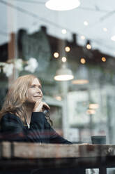 Thoughtful female professional sitting with hand on chin in cafe seen through glass window - JOSEF05178