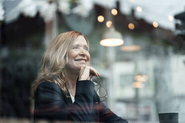 Glückliche blonde Frau, die mit der Hand am Kinn sitzt, gesehen durch ein Glasfenster - JOSEF05179