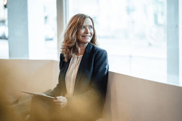 Smiling senior businesswoman with digital tablet looking through window while sitting in cafe - JOSEF05182