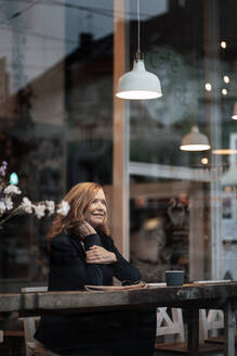 Lächelnde weibliche Fachkraft sitzt am Tisch in einem Café, gesehen durch ein Glasfenster - JOSEF05189