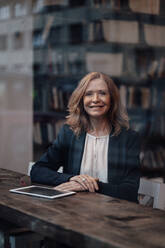 Lächelnde blonde Geschäftsfrau sitzt mit digitalem Tablet in einem Café, gesehen durch ein Glasfenster - JOSEF05194