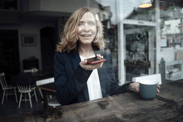 Smiling senior businesswoman holding coffee cup while talking on smart phone through speaker sitting in cafe - JOSEF05223
