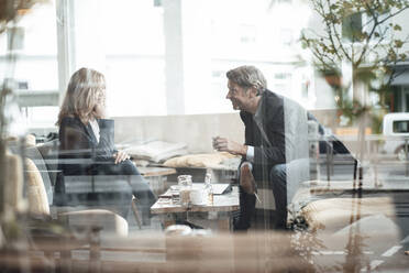 Smiling male and female colleagues sharing ideas sitting together in cafe seen through glass - JOSEF05228