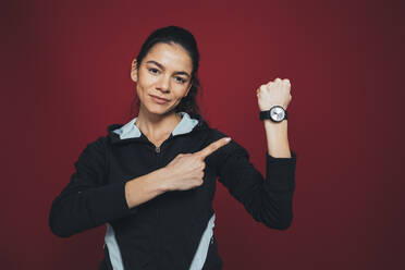 Mid adult female sportsperson pointing at her wristwatch against maroon background - OYF00393