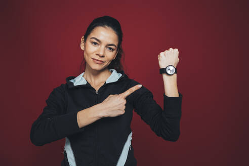 Mittlere erwachsene Sportlerin, die auf ihre Armbanduhr zeigt, vor kastanienbraunem Hintergrund - OYF00393