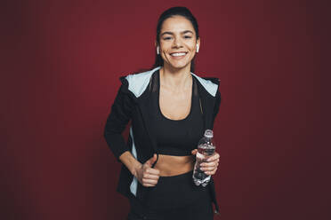 Lächelnde Sportlerin in Sportkleidung mit Wasserflasche vor kastanienbraunem Hintergrund - OYF00394