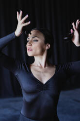 Young graceful woman in black apparel with raised arms playing castanets while performing traditional Spanish dance and looking away - ADSF27652