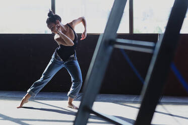 Junge barfüßige Frau in Jeans mit Haarknoten tanzt, während sie auf den Boden mit Schatten im Sonnenlicht schaut - ADSF27654