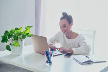 Cheerful female remote employee surfing internet on netbook at table with smartphone and copybook at home in sunlight - ADSF27660