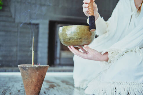 Crop woman playing singing bowl with wooden striker during spiritual practice - ADSF27663