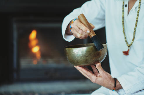 Crop man playing singing bowl with wooden striker during spiritual practice - ADSF27665