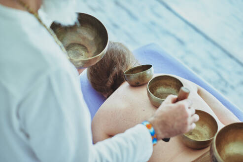 From above of crop unrecognizable male spiritual practitioner playing Himalayan bowls on back of topless female - ADSF27666