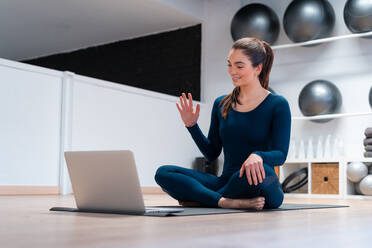 Ganzkörperansicht einer positiven jungen Lehrerin, die mit der Hand winkt, während sie die Schüler per Videochat auf dem Laptop während eines Online-Yoga-Kurses begrüßt - ADSF27679