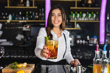 Glückliche junge Barkeeperin in stilvollem Outfit schaut in die Kamera und serviert Mojito-Cocktail mit Zitronenscheiben, während sie am Tresen einer modernen Bar steht - ADSF27686