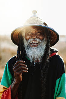 Portrait of cheerful old ethnic rastafari with dreadlocks looking at the camera in the nature with white background - ADSF27698