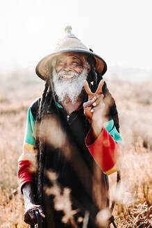 Fröhlicher alter ethnischer Rastafari mit Dreadlocks, der in die Kamera schaut, während er auf einer trockenen Wiese in der Natur steht - ADSF27705