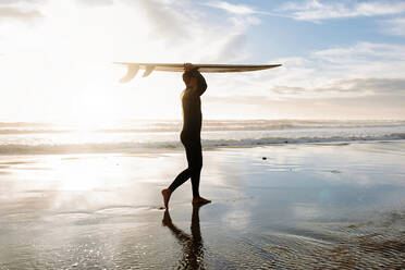 Seitenansicht von Surfer Mann in Neoprenanzug zu Fuß am Strand mit dem Surfbrett über dem Kopf in den Morgen mit Sonnenaufgang im Hintergrund gekleidet - ADSF27706