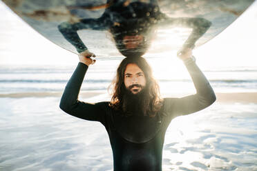 Porträt eines jungen Surfers mit langen Haaren und Bart in einem Neoprenanzug, der am Strand mit einem Surfbrett über dem Kopf bei Sonnenaufgang in die Kamera schaut - ADSF27707