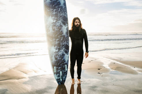 Surfer Mann mit langen Haaren und Bart in Neoprenanzug stehend Blick auf die Kamera mit dem Surfbrett am Strand während Sonnenaufgang im Hintergrund - ADSF27708