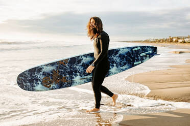 Seitenansicht eines Surfers im Neoprenanzug, der mit einem Surfbrett zum Wasser läuft, um eine Welle am Strand bei Sonnenaufgang zu erwischen - ADSF27709