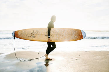Seitenansicht von Surfer Mann in Neoprenanzug zu Fuß am Strand mit dem Surfbrett am Morgen mit Sonnenaufgang im Hintergrund gekleidet - ADSF27711