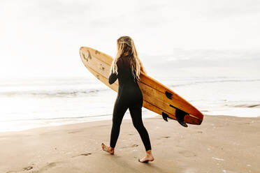 Seitenansicht einer Surferin im Neoprenanzug, die mit einem Surfbrett über dem Kopf am Strand spazieren geht, während im Hintergrund die Sonne aufgeht - ADSF27712