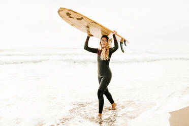 Seitenansicht einer weiblichen Surferin im Neoprenanzug, die am Strand bei Sonnenaufgang im Hintergrund ein Surfbrett auf dem Kopf hält und dabei wegschaut - ADSF27716