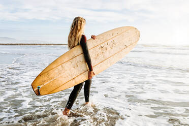 Seitenansicht einer Surferin im Neoprenanzug, die mit einem Surfbrett über dem Kopf am Strand spazieren geht, während im Hintergrund die Sonne aufgeht - ADSF27717