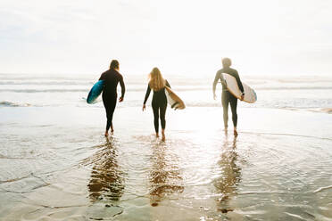 Rückenansicht einer nicht erkennbaren Gruppe von Surferfreunden in Neoprenanzügen, die mit Surfbrettern zum Wasser laufen, um am Strand bei Sonnenaufgang eine Welle zu erwischen - ADSF27718