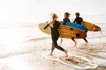 Seitenansicht einer Gruppe von Surferfreunden in Neoprenanzügen, die mit Surfbrettern zum Wasser laufen, um eine Welle am Strand bei Sonnenaufgang zu erwischen - ADSF27719