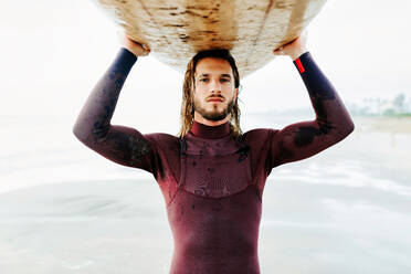 Porträt eines jungen Surfers mit langen Haaren und Bart in einem Neoprenanzug, der am Strand mit einem Surfbrett über dem Kopf bei Sonnenaufgang in die Kamera schaut - ADSF27720