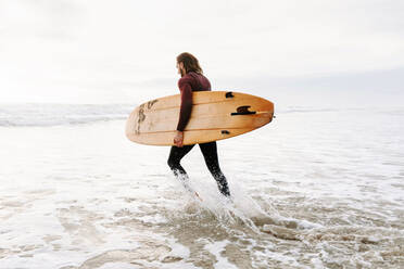 Seitenansicht eines Surfers im Neoprenanzug, der mit einem Surfbrett zum Wasser läuft, um eine Welle am Strand bei Sonnenaufgang zu erwischen - ADSF27722